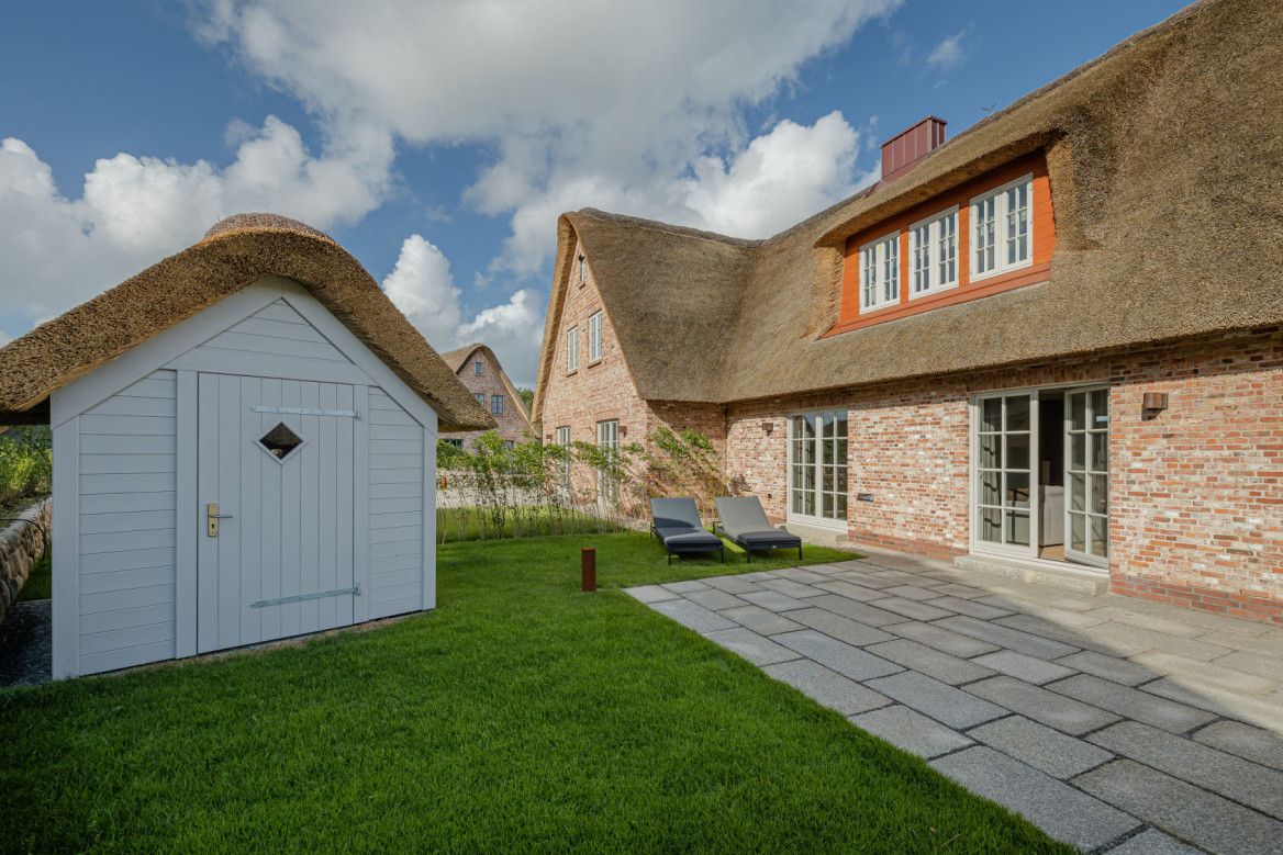 Ebenerdiger Zugang Reetgedecktes Haus mit Garten und kleiner weißer Gartenhütte unter bewölktem Himmel.