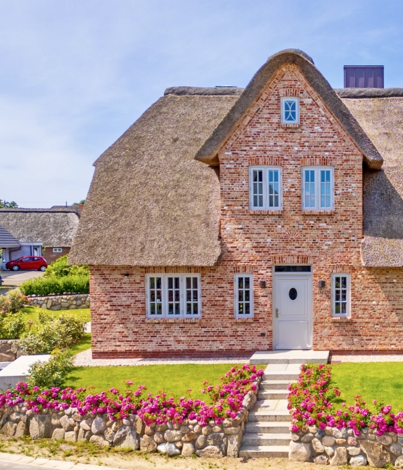 Vom Parkplatz zur Tür. Zweistöckiges, rotes Backsteinhaus mit Reetdach und blühenden Blumen im Vordergrund.